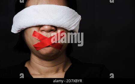 Slave Asian woman blindfold wrapping mouth with red adhesive tape, tied with chains and closed her eyes black background. Freedom speech censorship an Stock Photo