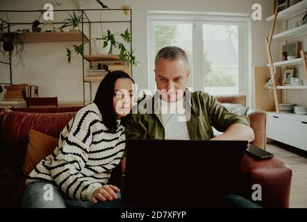 Serious husband and wife using laptop at home browsing while sitting together on sofa Stock Photo