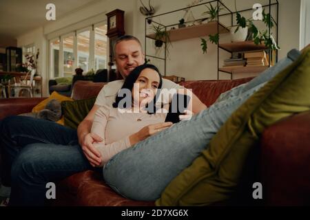Smiling young woman using phone while relaxing with husband on couch at home Stock Photo