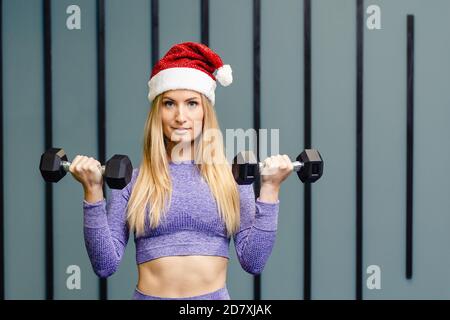 Sporty girl in the Santa Claus hat raises weights on grey background. Christmas, holidays, fitness, and gym concept. Copy space. Stock Photo