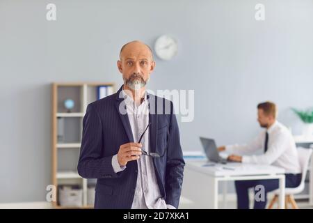 Mature company executive standing in office looking at camera with employee working in background Stock Photo