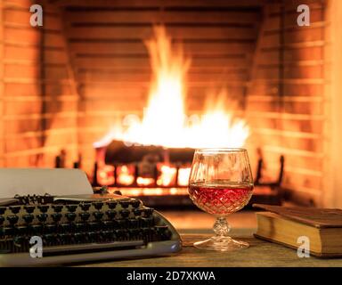 Glass of brandy and a typewriter on burning fireplace background. Warm cozy author home interior Stock Photo