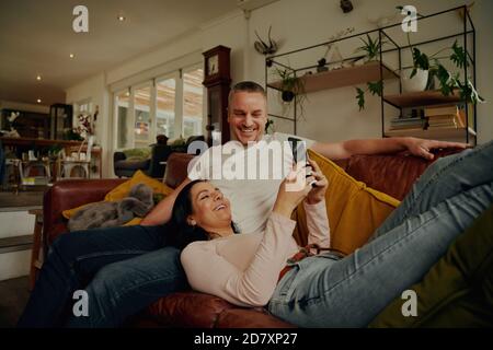 Cheerful young wife using phone while lying on husband's lap and relaxing on couch at home Stock Photo
