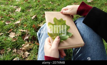 A woman holds a closed book lying in her lap with a fallen oak leaf close-up in a park on a sunny warm autumn day. The concept of relaxation, reading Stock Photo