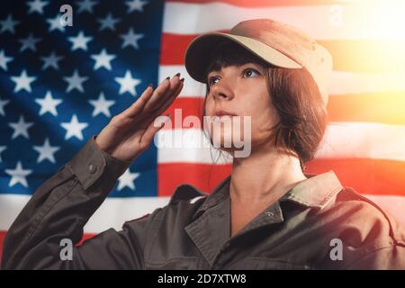 USA celebration. Portrait of a female soldier saluting, against the background of the American flag. Side view. Light. Stock Photo