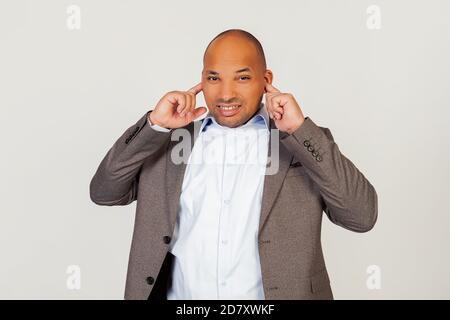 Portrait of unhappy young african american guy businessman covering ears with fingers with annoyed facial expression trying not to hear bad singing and no pleasant sound. Standing on a gray background Stock Photo
