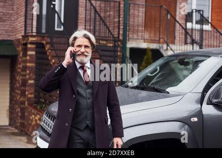 Fashionable senior man with gray hair and beard is outdoors on the street near his car have conversation by the phone
