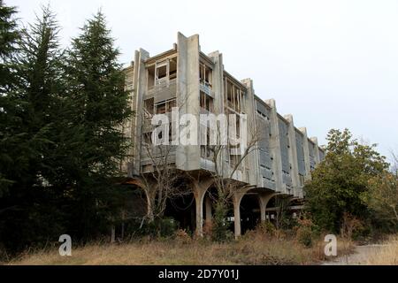 Cold War era abandoned hotel complex with destroyed facade and windows on top of small hill surrounded with tall trees and dry uncut grass Stock Photo