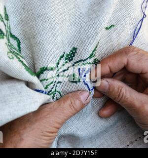 Hands of an elderly woman embroidering a cross-stitch floral pattern on linen fabric. Embroidery, handwork, needlecraft concept. Close-up Stock Photo