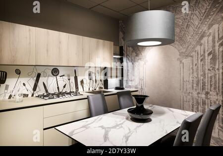 Modern fitted kitchen with decorative splash back and wall with a dining table and chairs under a hanging ceiling light illuminating the room Stock Photo