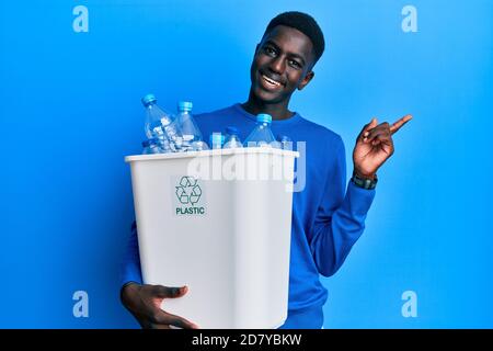 Young african american man holding recycling wastebasket with plastic bottles smiling happy pointing with hand and finger to the side Stock Photo
