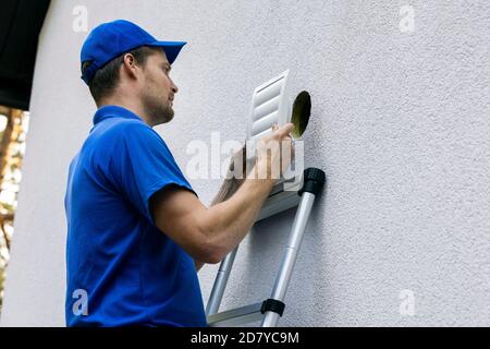 home ventilation - hvac worker standing on ladder and installing air duct grille on house facade Stock Photo