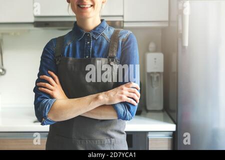 young smiling woman with apron standing in home kitchen with arms crossed Stock Photo