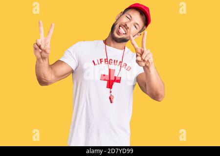 Young caucasian man wearing lifeguard t shirt holding whistle smiling with tongue out showing fingers of both hands doing victory sign. number two. Stock Photo