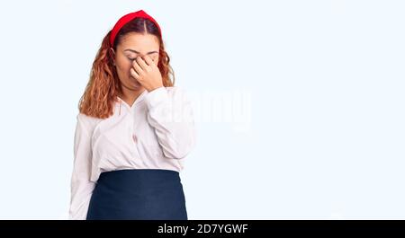 Young latin woman wearing waitress apron tired rubbing nose and eyes feeling fatigue and headache. stress and frustration concept. Stock Photo