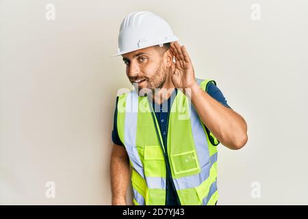 Handsome man with beard wearing safety helmet and reflective jacket smiling with hand over ear listening and hearing to rumor or gossip. deafness conc Stock Photo