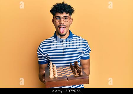 Young arab handsome man holding chess board wearing glasses sticking tongue out happy with funny expression. Stock Photo