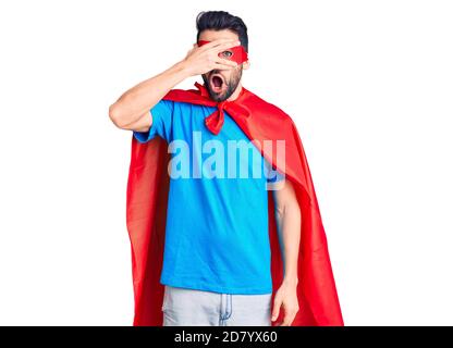 Young handsome man with beard wearing super hero costume peeking in shock covering face and eyes with hand, looking through fingers with embarrassed e Stock Photo