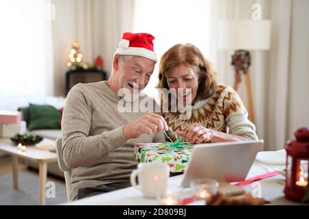 Senior couple opening present indoors at home at Christmas, having video call with family. Stock Photo
