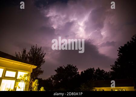 Bolt of lightning on a summer night lights up the sky purple. Corner of a house lit in the foreground as the flash happens above a distant treeline Stock Photo