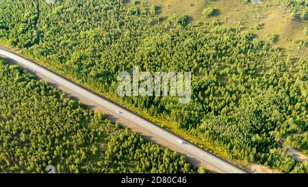 Aerial flying over a rural road with two cars driving opposite each other on the lane between forest on a sunny day. Scenic bird eye view from drone o Stock Photo