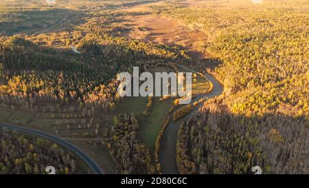 Amazing aerial panorama from drone over a river, fall forest, hill and asphalt road. Beautiful autumn nature rural landscape on sunset from birds eye Stock Photo