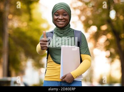 Black Muslim Female Student In Hijab Posing Outdoors, Showing Thumb Up Stock Photo