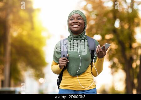 Black muslim woman in hijab listening music with smartphone and earphones outdoors Stock Photo