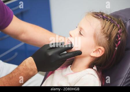 Portrait of little girl has a dental examination while sitting at dentist office. Male dentist is checking the young patient's mouth. Stock Photo