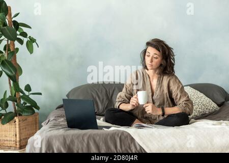 A young brunette woman uses a laptop in her bedroom while sitting on the sofa. Stock Photo