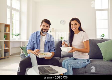 Man and woman holding takeaway food and having lunch at home sitting on sofa in front of laptop. Stock Photo