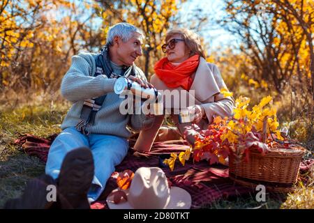 Senior family couple having tea pouring it from thermos in fall forest. Happy man and woman enjoying picnic outdoors. Stock Photo