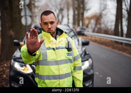 Male police officer in green uniform showing stop gesture near vehicle Stock Photo