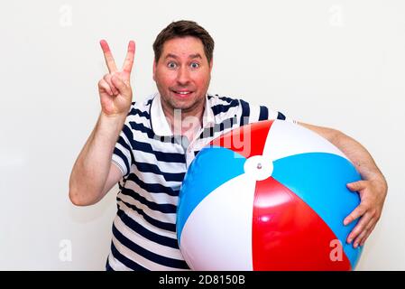 portrait of a surprised excited middle age guy holding beach ball and showing victory gesture on white studio wall. Stock Photo
