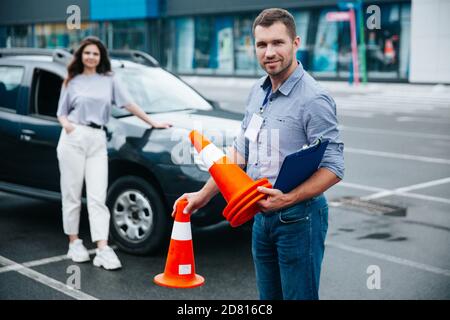 Happy male driving instructor holding traffic cones in his hands, looking at camera and smiling. Young woman standing near the car and waiting when ma Stock Photo