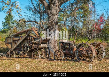 An old antique harvester and tractor no longer in use rusty and weathered abandoned in a farm field next to a large old tree on a bright sunny day in Stock Photo