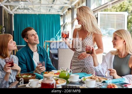 blonde caucasian woman say a toast in honor of the birthday of friend or colleague in restaurant, they drink beverages and eat delicious tasty food, meal Stock Photo