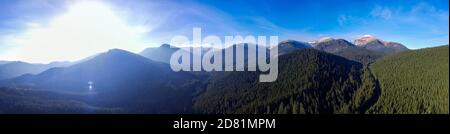Bright sun rising over the mountains. Panorama of mountain landscape and dense forest from aerial view. Stock Photo