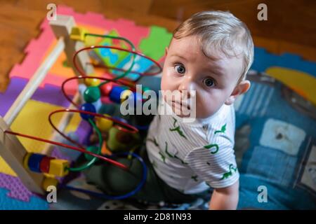 Portrait of creative little curious baby boy sitting on small cushion in living room playing with wooden bead maze game at home Stock Photo