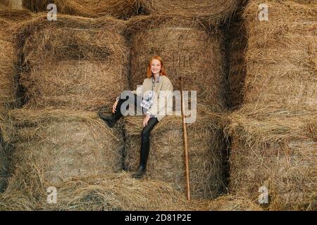 Ginger girl sitting on piled up hay bundles inside a barn Stock Photo