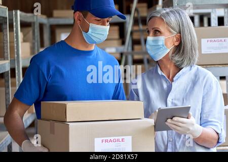 Female manager and courier wearing face masks talking in warehouse. Stock Photo