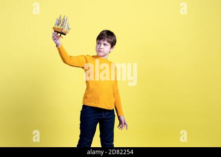 A 9-year-old boy plays with a handmade toy boat and imagines himself as a captain and a traveler on a yellow background in the Studio.  Stock Photo