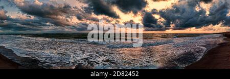 Dramatic weather panorama over the sea threatening waves crashing at the shore with overcast golden sky with beautiful sunbeams reflections between he Stock Photo