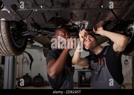 two interracial diverse mechanics checking car bottom in auto service, cooperate, work together Stock Photo