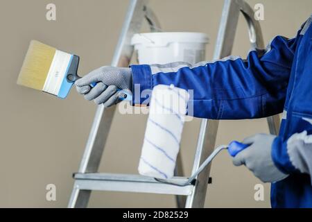 The plaster painter is ready to paint the wall. In the hands of a roller and brush. A stepladder and a bucket of paint in the background Stock Photo