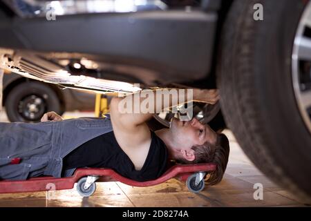 side view on concentrated auto mechanic male working alone on the floor repairing bottom of the car, look at details, troubleshoot the car Stock Photo