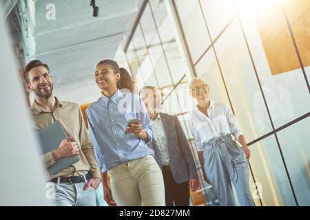 Two young happy diverse male and female coworkers discussing something and smiling while walking with colleagues in the modern office or coworking Stock Photo
