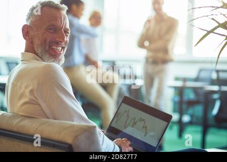 Young cheerful mature businessman sitting in the modern office, using laptop and smiling at camera while his colleagues talking on the background Stock Photo