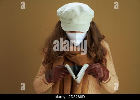 Life during coronavirus pandemic. unhappy elegant middle aged woman in sweater with white medical mask and wallet isolated on brown background. Stock Photo
