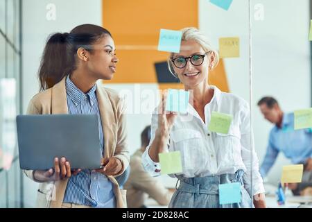 wo happy female coworkers putting colorful sticky notes on a glass wall and smiling while standing in the modern office, business people working Stock Photo